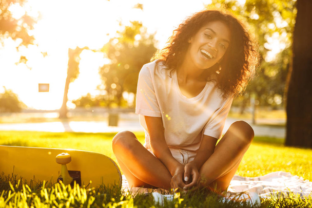 Image of happy young african woman on grass outdoors in park near skateboard. - Zdjęcie, obraz