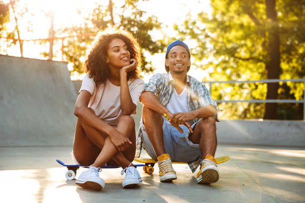 Portrait of a happy young african couple with skateboard sitting together at the skate park - Photo, Image