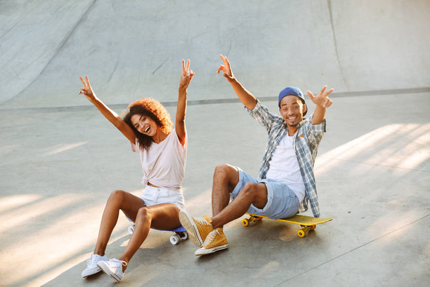 Portrait of a happy young african couple with skateboards sitting together at the skate park - Zdjęcie, obraz