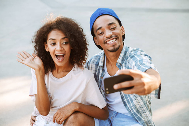 Portrait of a happy young african couple taking a selfie together at the skate park, waving - Photo, image