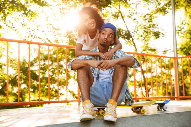 Portrait of a joyful young african couple with skateboards sitting together at the skate park - Foto, Imagem