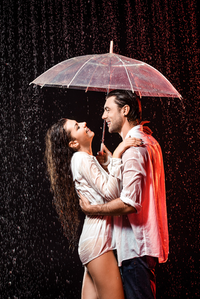 Zijaanzicht van romantische echtpaar in witte shirts met paraplu staan onder regen op zwarte achtergrond - Foto, afbeelding