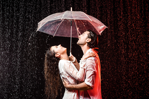 vista lateral de pareja romántica en camisas blancas con paraguas de pie bajo la lluvia sobre fondo negro
 - Foto, Imagen