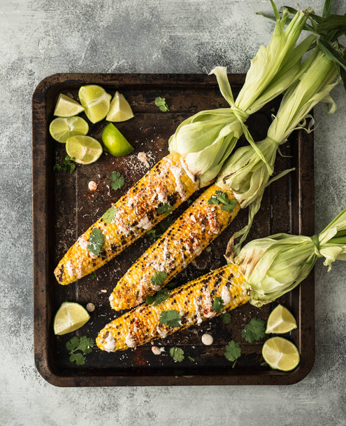 Grilled corn cobs with sauce, coriander, lime, paprika and cheese on metal baking tray. Mexican food. Top view - Fotó, kép