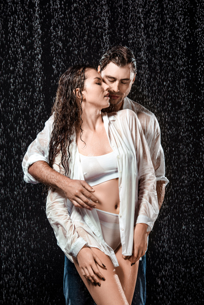 joven sexy pareja en blanco camisas de pie bajo lluvia aislado en negro
 - Foto, Imagen