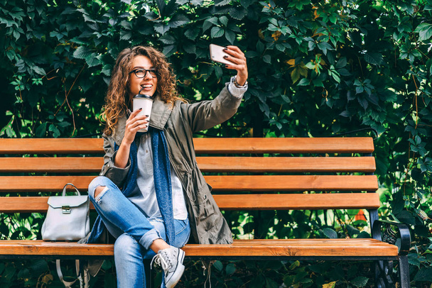 Pretty girl with curly hair drinking coffee and making selfie on smartphone outdoor - Фото, изображение