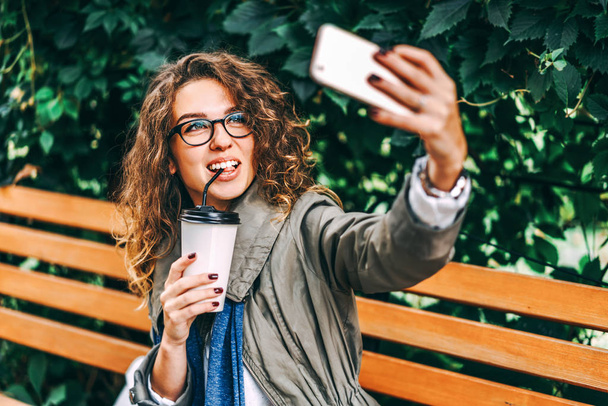 Pretty girl with curly hair drinking coffee and making selfie on smartphone outdoor - Foto, afbeelding