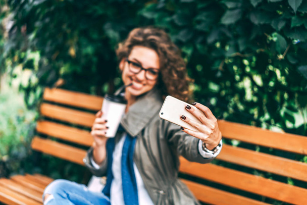 Pretty girl with curly hair drinking coffee and making selfie on smartphone outdoor - Fotó, kép
