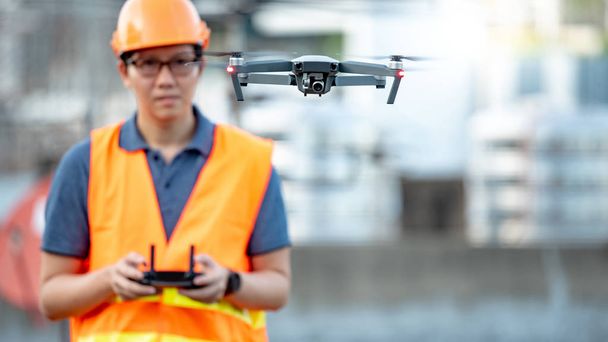 Joven ingeniero asiático volando drone sobre el sitio de construcción. Uso de vehículos aéreos no tripulados (UAV) para el estudio de terrenos y obras en proyectos de ingeniería civil
. - Foto, Imagen