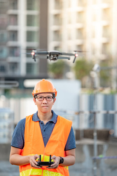 Jovem engenheiro asiático voando drone sobre canteiro de obras. Utilização de veículos aéreos não tripulados (VANT) para o levantamento de terrenos e locais de construção em projectos de engenharia civil
. - Foto, Imagem