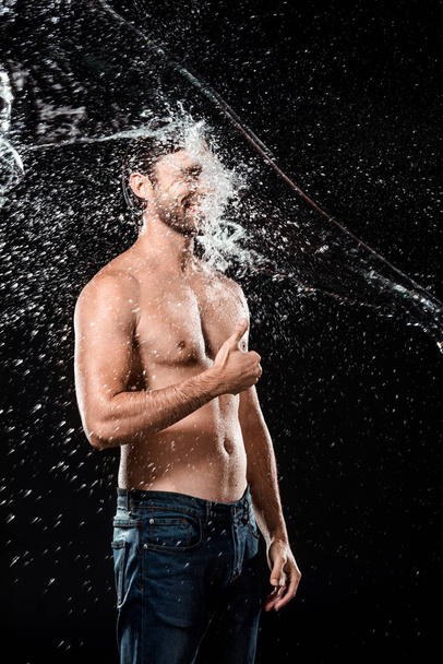 smiling young man showing thumb up while swilled with water isolated on black - Photo, Image