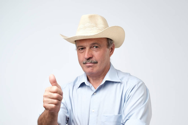 Hombre maduro guapo en sombrero de vaquero sonriendo en la cámara con el pulgar hacia arriba aprobando su elección. Estudio rodaje sobre fondo gris
 - Foto, Imagen