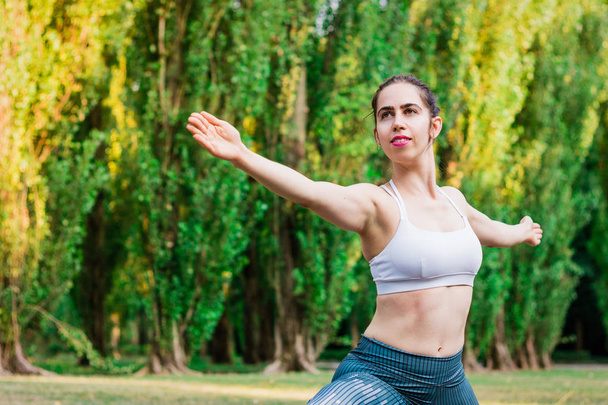 Slim young woman practicing yoga in nature, standing in Warrior Two exercise, Virabhadrasana II pose. Green background. Good healthy and sport concept.  - Photo, Image