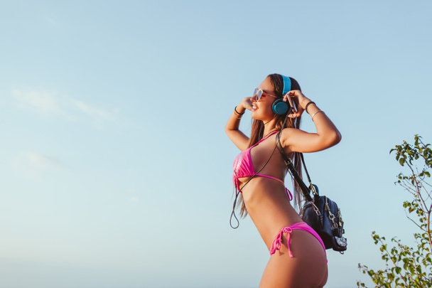 Seitenansicht eines attraktiven Mädchens mit Kopfhörern und rosa Bikini, das vor blauem Himmel posiert - Foto, Bild