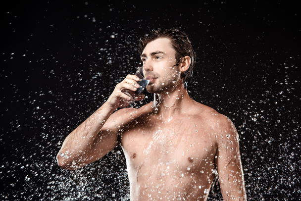 portrait of shirtless man drinking water from glass while swilled with water isolated on black - Zdjęcie, obraz