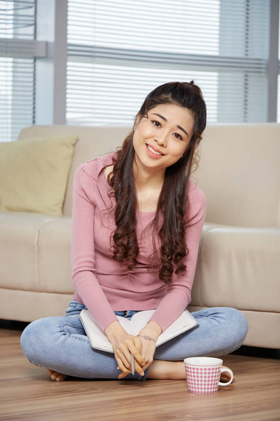 Atractiva joven asiática sentada en el suelo en la habitación en el sofá y sosteniendo el libro de rodillas y mirando a la cámara
  - Foto, Imagen