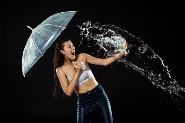 young emotional woman with umbrella swilled with water isolated on black - Fotó, kép