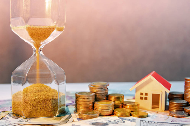 Saving money for retirement concept. Real estate or property investment. Home mortgage loan rate. Gold coin stack, house model and hourglasses on international banknotes. - Photo, Image