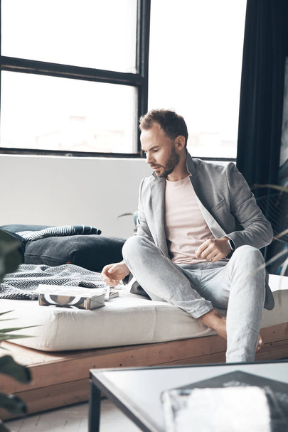 Handsome man with dark hair and beard in light jacket and linen pants sits on the bed in scandinavian studio with big window and plants - Zdjęcie, obraz