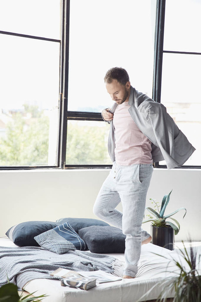 Handsome man with dark hair and beard in light jacket and linen pants stands on the bed in scandinavian studio with big window and plants - Foto, afbeelding