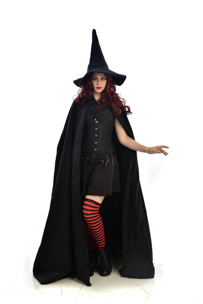 Ολόσωμο πορτρέτο της κόκκινα μαλλιά κορίτσι φορώντας μακρύ μαύρο μανδύα, μυτερό καπέλο και κοστούμι μάγισσα. όρθια στάση, απομονώνονται σε φόντο λευκό στούντιο. - Φωτογραφία, εικόνα