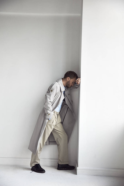 Fashion portrait of handsome man with dark beard and hair, weared in light trench coat, shirt, beige pants and black shoes. Man facing white wall - Фото, изображение