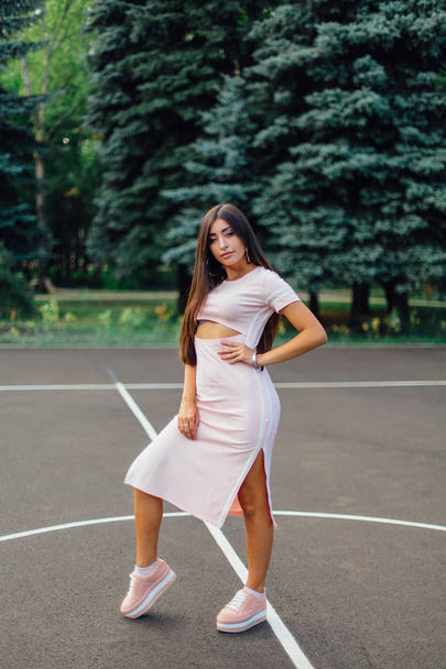 Charming brunette female dressed in a pink dress posing on the basketball court during sunset. - Фото, зображення