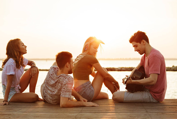 Image of young happy friends sitting outdoors on the beach play on the guitar drinking beer. - Photo, image