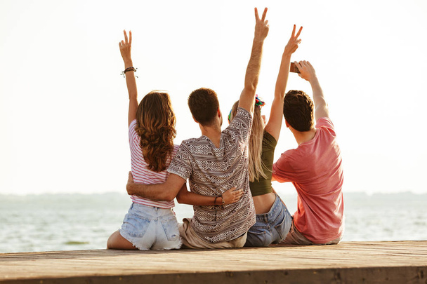 Picture of group of four friends loving couples walking outdoors on the beach showing peace gesture make a selfie. - Φωτογραφία, εικόνα