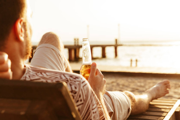Foto de un hombre guapo descansando al aire libre en la playa bebiendo cerveza
. - Foto, Imagen