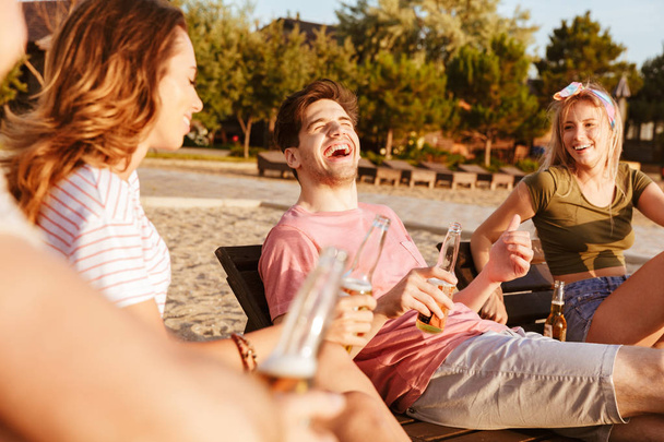 Group of cheerful friends dressed in summer clothing drinking beer while relaxing at the beach - Foto, Imagen
