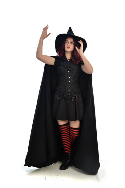 retrato de comprimento total da menina de cabelos vermelhos vestindo manto preto longo, chapéu pontiagudo e traje de bruxa. em pé pose, isolado no fundo do estúdio branco
. - Foto, Imagem