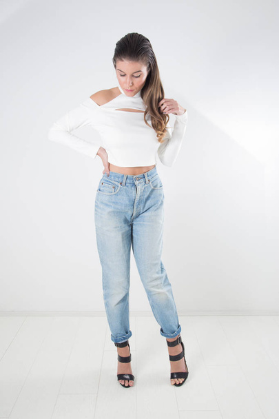 Stylish Young Woman With Long Slicked Back Hair Wearing Jeans, White Blouse With Bare Shoulders And Black High Heeled Sandals With Straps Posing At Studio - Foto, imagen