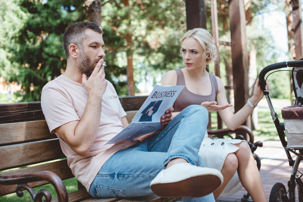 manžel kouří cigaretu a čtenářský deník poblíž kočárek v parku, při pohledu na něj naštvaná manželka - Fotografie, Obrázek