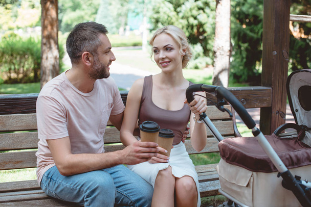 a szülők közelében babakocsi parkban a padon ül, és a gazdaság kávé papírpohár - Fotó, kép