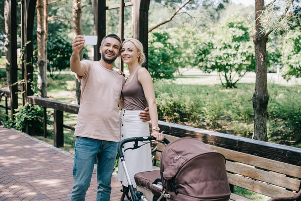 μητέρα και ο πατέρας λαμβάνοντας αυτοπορτρέτα με έξυπνο κινητό τηλέφωνο κοντά σε baby μεταφορά στο πάρκο - Φωτογραφία, εικόνα