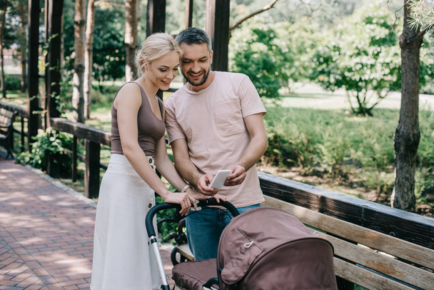 parents taking photo of baby with smartphone near baby carriage in park - Foto, imagen