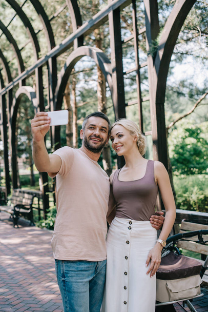 mosolygó szülők vesz smartphone közelében babakocsi selfie park - Fotó, kép