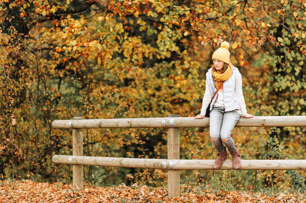 Осенний портрет очаровательной девочки-подростка 10-11 лет, в сером пуховом жилете, наборе желтой шляпы и шарфа, сидящей на заборе
 - Фото, изображение