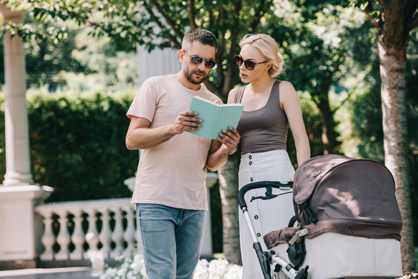 parents standing near baby carriage in park and reading book - Fotoğraf, Görsel