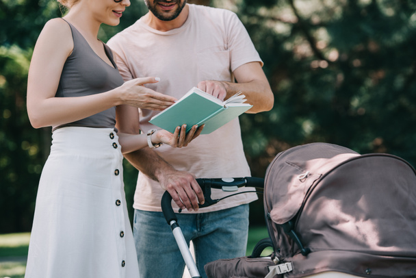 περικομμένη εικόνα των γονέων στέκεται κοντά baby μεταφορά στο πάρκο και την ανάγνωση του βιβλίου - Φωτογραφία, εικόνα