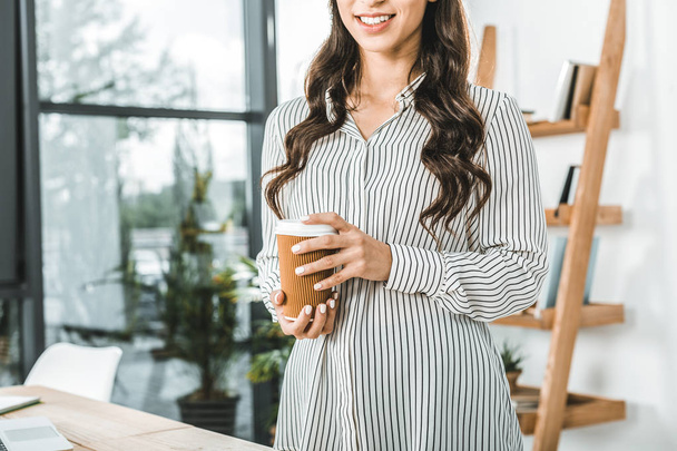 partial view of smiling businesswoman holding coffee to go in hands in office - Photo, image