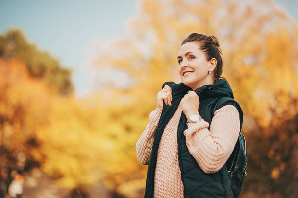Outdoor autumn portrait of young happy woman wearing warm pullover and black down vest, yellow foliage on background - Photo, image