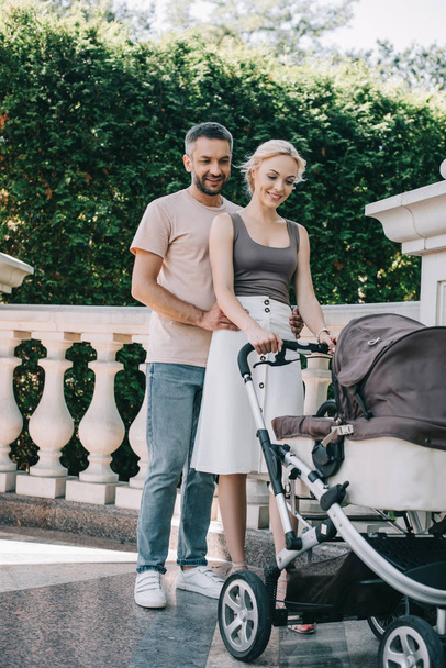 husband hugging wife near with baby carriage in park and they looking at kid - Photo, Image