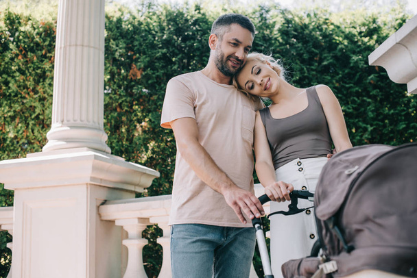 smiling parents cuddling and looking at baby carriage in park - Photo, Image
