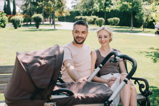 happy parents sitting on bench near baby carriage in park - Foto, Imagen