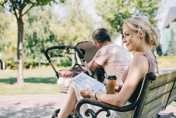 side view of wife sitting near baby carriage with coffee to go and travel newspaper - Fotografie, Obrázek
