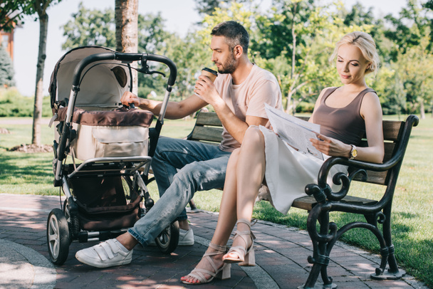 padre bebiendo café y tocando carro de bebé en el parque, madre leyendo periódico
 - Foto, Imagen