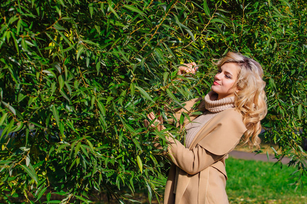 Beautiful elegant blonde woman dressed in a coat standing under the tree in autumn park. Sunny october day. - Foto, afbeelding