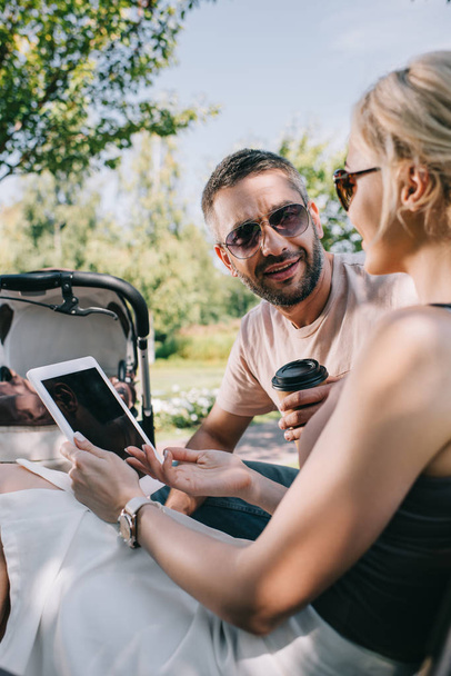 parents sitting near baby carriage in park and holding tablet with blank screen - Фото, изображение